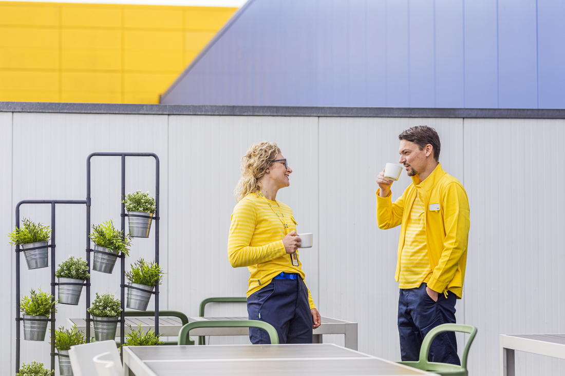 Two IKEA coworkers in yellow uniforms chatting and drinking coffee on an outdoor terrace.