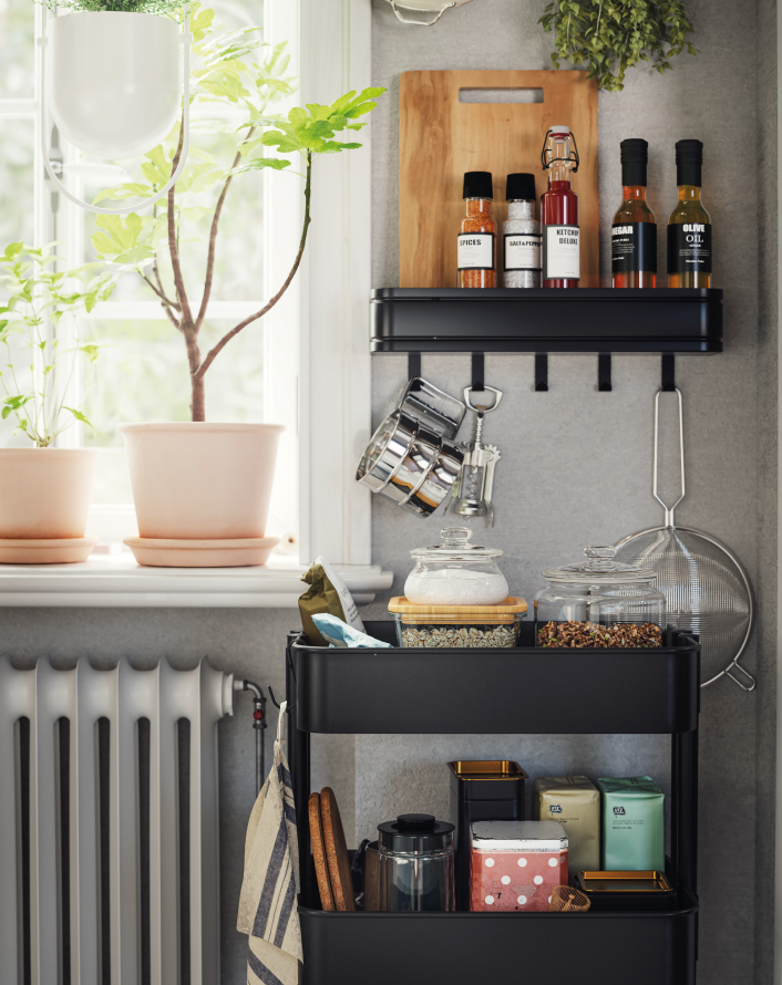 Black kitchen shelf with spices, jars, oils, and utensils next to a window with plants.