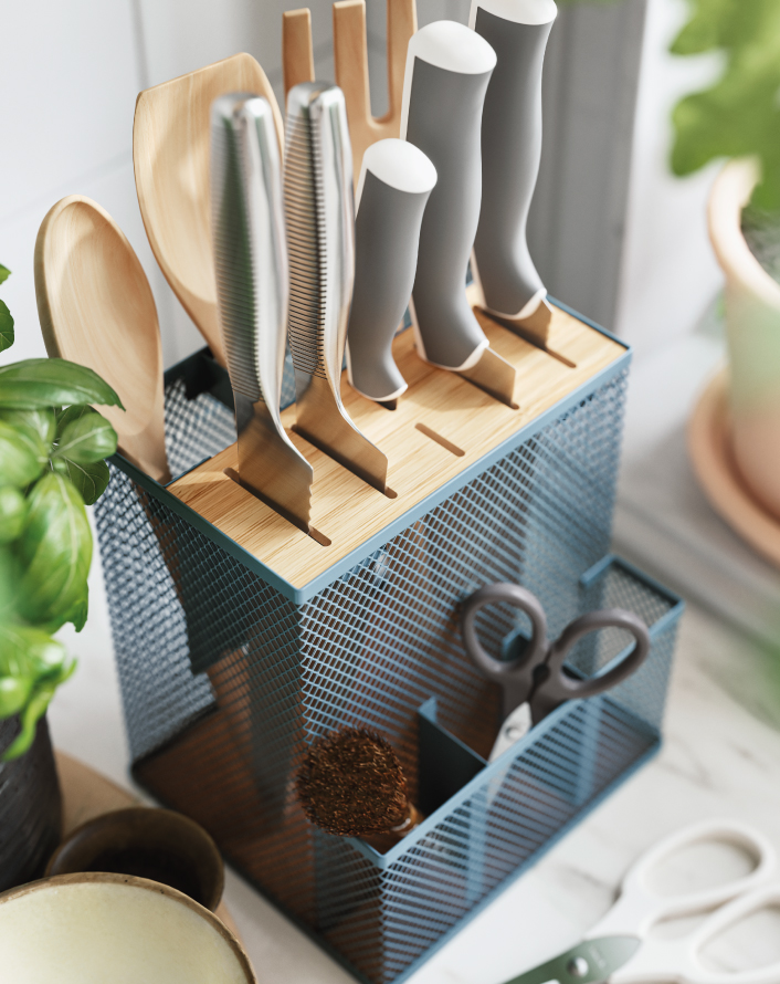 Kitchen organizer with knives, utensils, and scissors in a metal stand on a countertop next to plants.