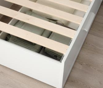 High-angle view of a white bed frame with wooden slats, showing storage space underneath filled with folded green blankets.