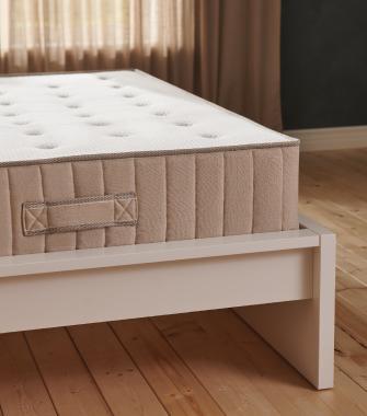 Close-up of a thick beige mattress with a side handle, resting on a minimalist white wooden bed frame.