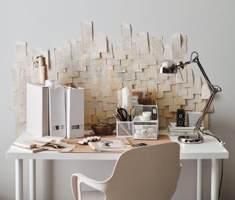 White home office desk with a silver lamp, organizers, and a large artistic collage of paper scraps on the wall behind it.
