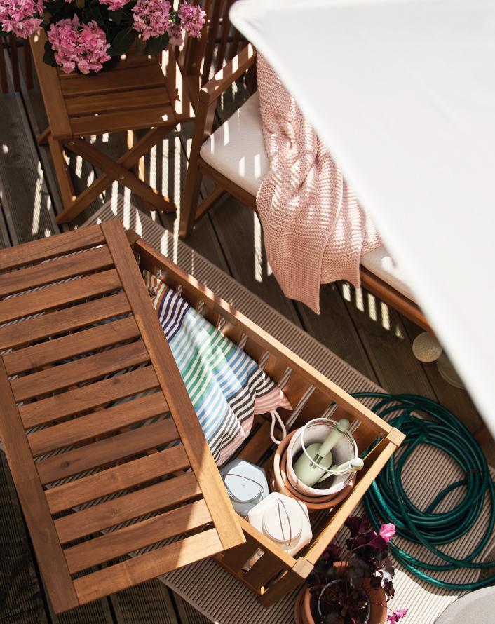 Outdoor wooden storage bench with open compartment holding cushions, watering cans, and garden accessories.