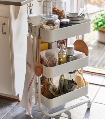Un carrito de cocina de tres niveles de color blanco hueso sobre ruedas, cargado con frascos de comida, toallas colgadas y utensilios, de pie sobre baldosas de piedra.