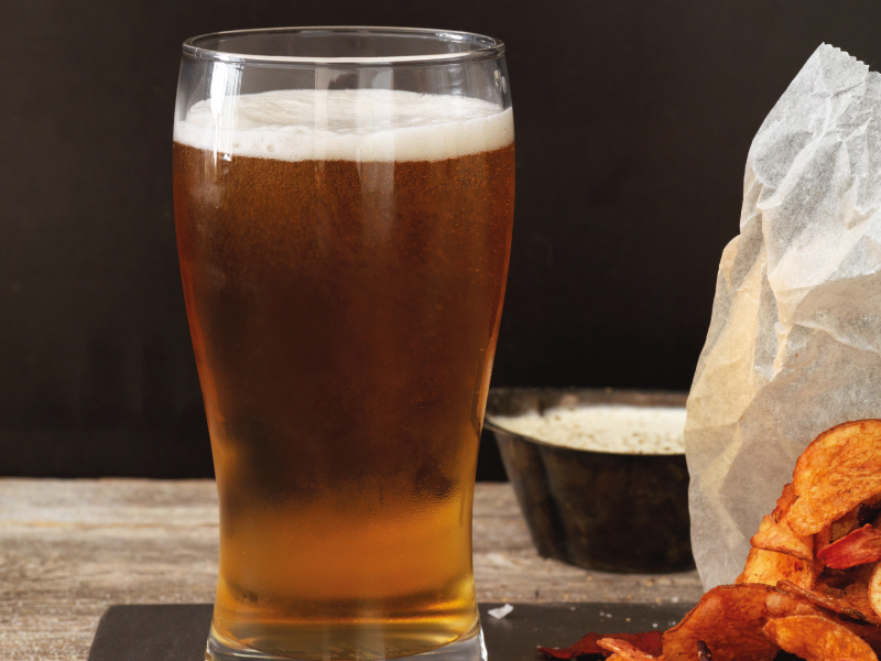 Glass of beer with foam next to potato chips.