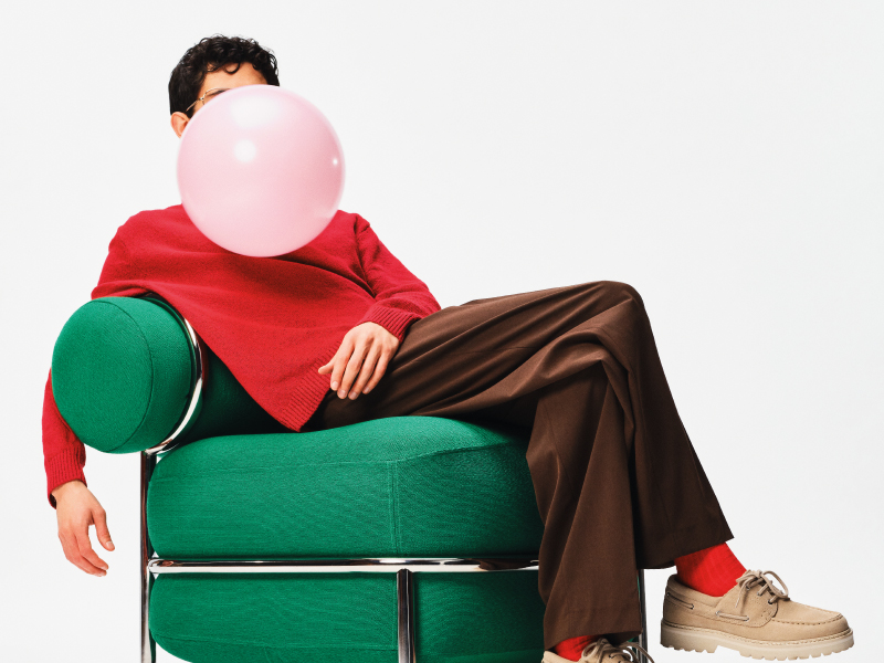 Person sitting on a green chair blowing a pink balloon.