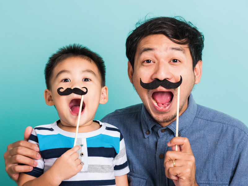 Happy father and young son holding fake black mustaches on sticks and yelling against a turquoise background.