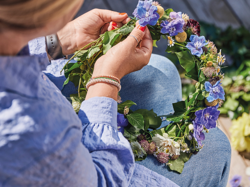 Close-up of a person's hands weaving a colorful flower crown with blue, white, and purple blossoms and green leaves.