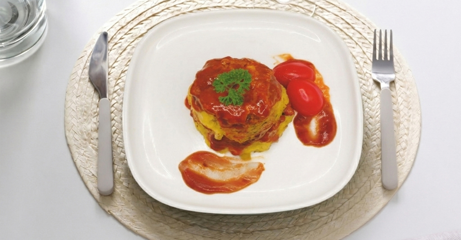 A top-down view of a round savory dish covered in red sauce and topped with a parsley sprig, served on a square white plate with two cherry tomatoes, a knife, and a fork.