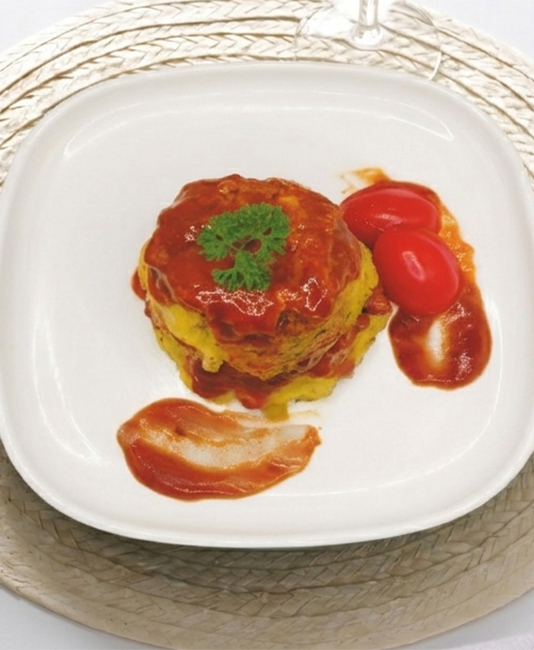 A top-down view of a round savory dish covered in red sauce and topped with a parsley sprig, served on a square white plate with two cherry tomatoes, a knife, and a fork.