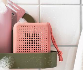 Altavoz portátil rosa con una correa tejida sobre un estante de baño.