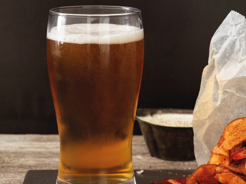 Glass of beer with foam next to potato chips.