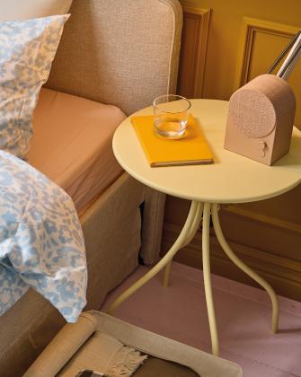 Mesa auxiliar redonda junto a una cama con un vaso, un cuaderno amarillo y un altavoz decorativo.