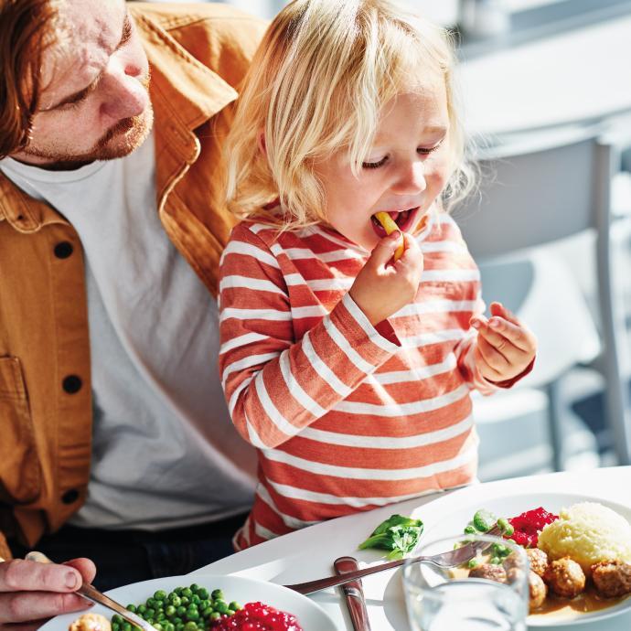 Un hombre mira con ternura a su pequeña hija rubia mientras ella come una patata frita sentada a la mesa con platos de comida.
