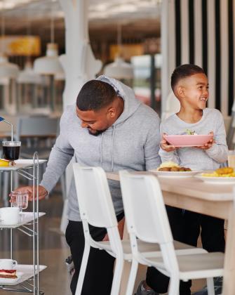 Un hombre con sudadera gris toma un vaso de un carrito mientras su hijo pequeño sonríe sosteniendo un plato rosa en un restaurante luminoso.
