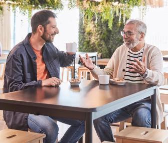 Un hombre joven y un adulto mayor conversan alegremente y gesticulan mientras toman café sentados a la mesa de una cafetería moderna.