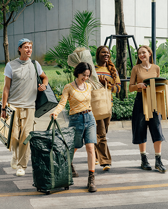 Grupo de personas caminando con bolsas, una planta y banquetas de madera.