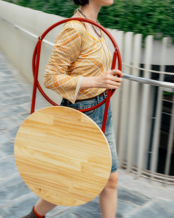 Persona caminando mientras lleva una mesa plegable redonda de madera con patas rojas.