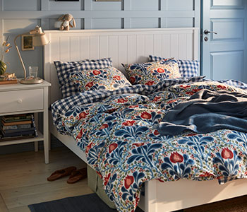 Un dormitorio pintado de azul con un juego de funda nórdica floral de la colección SÄTERVIDE sobre una cama blanca, junto a una mesita de noche blanca con una lámpara.