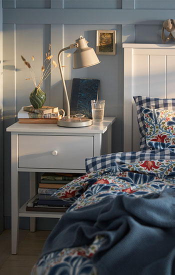 Una cama blanca con un juego de funda nórdica SÄTERVIDE con estampado floral multicolor junto a una mesita de noche blanca con una lámpara.