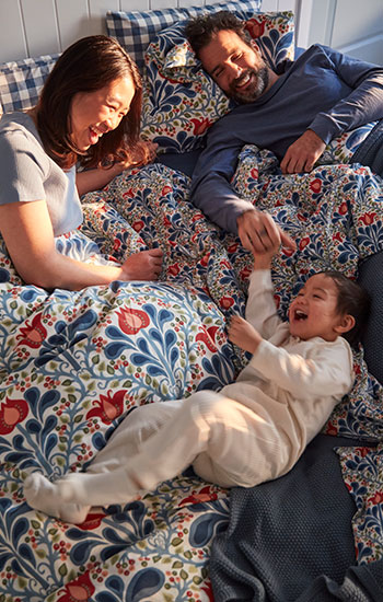 Una familia riendo en la cama con un juego de funda nórdica SÄTERVIDE con estampado floral multicolor en tonos naranja y azul.