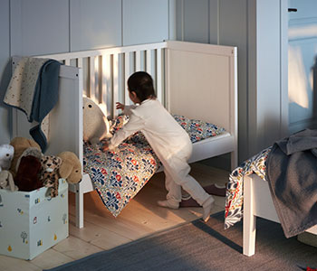 A child in white pyjamas is reaching for a soft toy inside a white SUNDVIK wooden cot with an open long side.