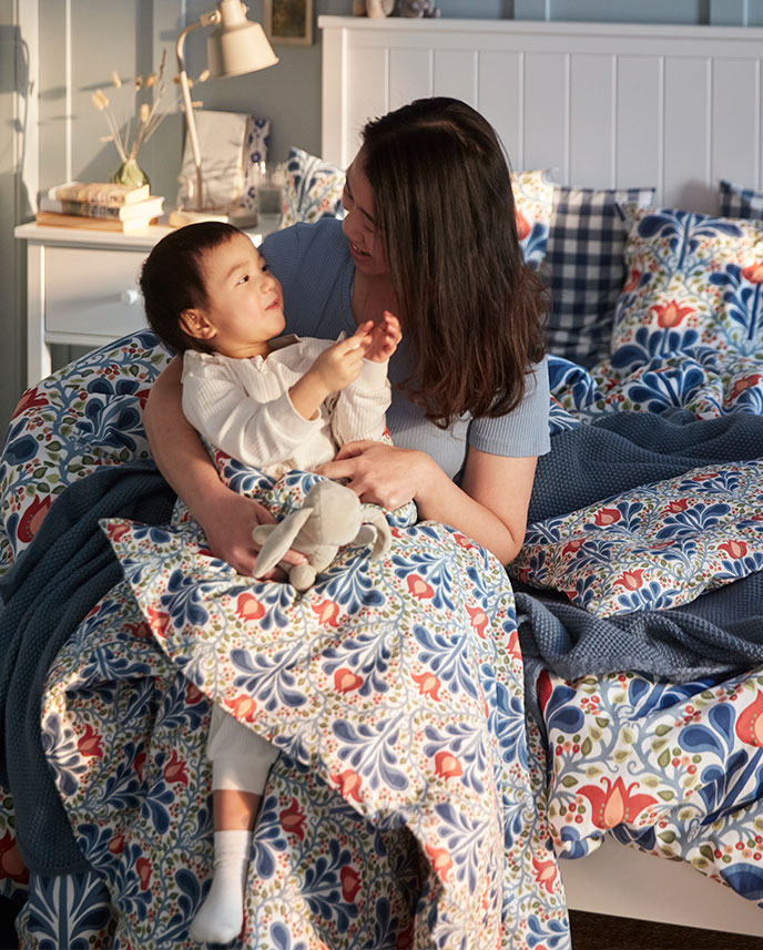 A person and a child sit on a bed with a multicolour SÄTERVIDE duvet cover and pillowcases in a floral pattern.