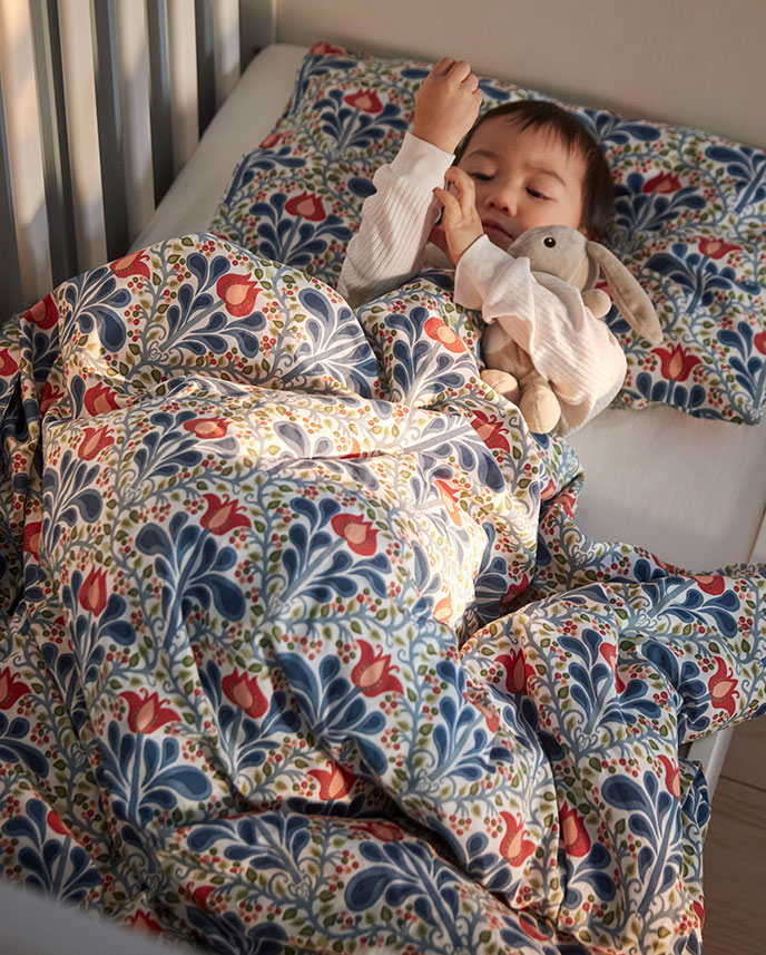 A child in white pyjamas is lying in a white SUNDVIK cot with a floral multicoloured SÄTERVIDE duvet cover set.
