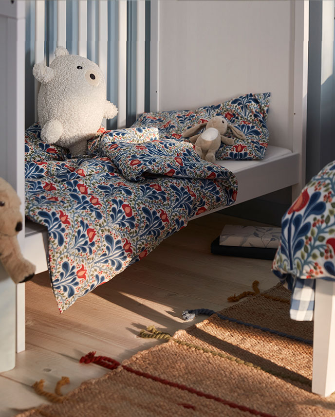A white SUNDVIK cot with one long side removed has a floral SÄTERVIDE duvet cover and pillowcase and two soft toys inside.