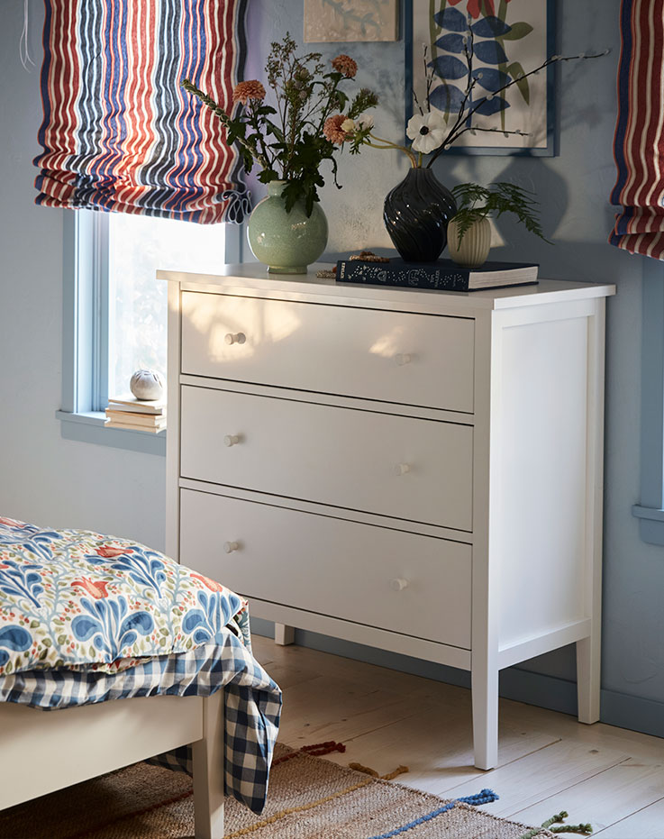A white GULLABERG chest of drawers stands by the end of a white bed in a blue bedroom with striped blinds.