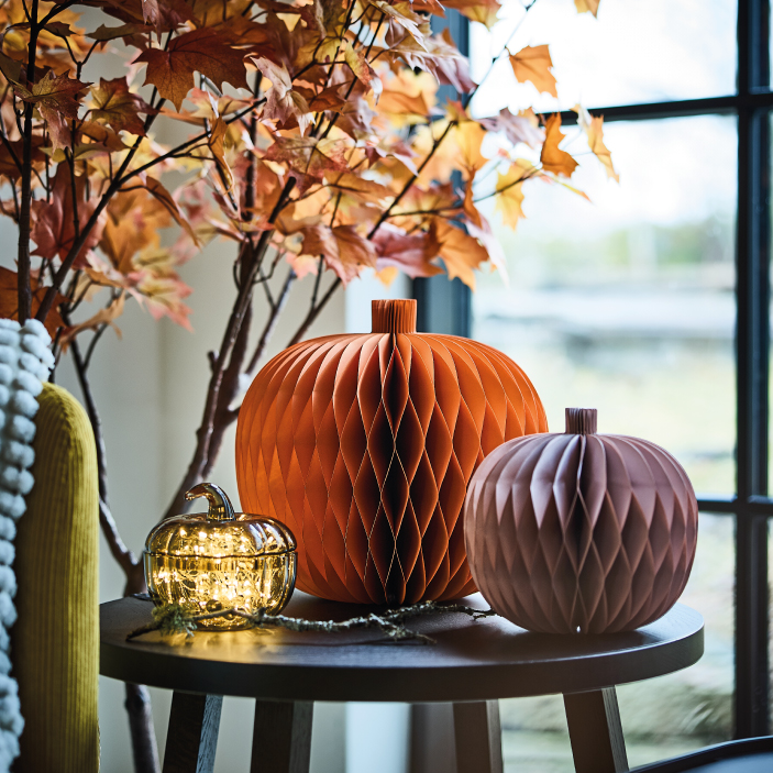 Decorative paper pumpkins on a table beside autumn leaves and a window.