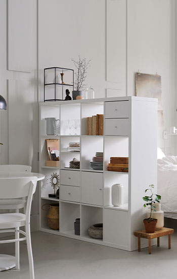 A bedroom featuring a white KALLAX shelving unit filled with decor and storage boxes next to a double bed with white linen.