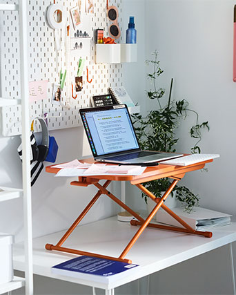 Soporte elevador naranja para portátil sobre un escritorio blanco con panel organizador de pared.