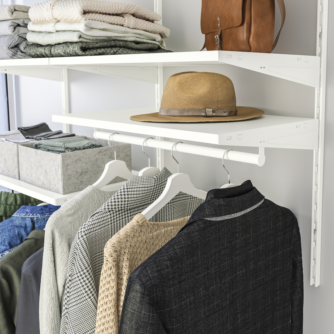 Sistema de estantería modular blanca de pared con ropa colgada en ganchos, suéteres doblados, un sombrero marrón y un bolso de cuero.